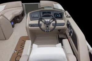 A steering area of a modern boat with tan upholstery. Features a steering wheel, digital dashboard, and control panel. The seating area is partially visible with beige cushions, and the boat flooring has a design element with the word "Avalon.