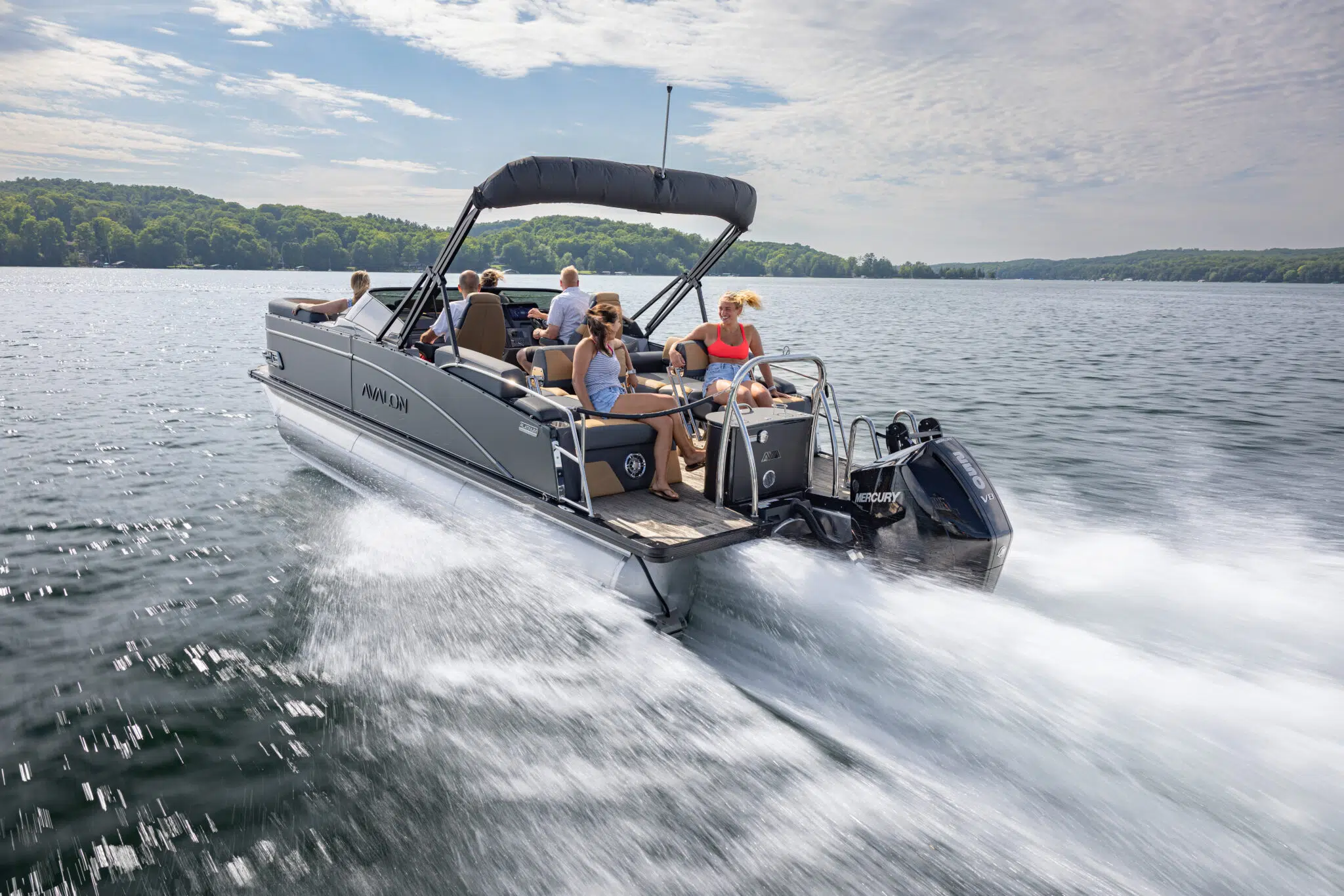 A group of people enjoying a ride on a Catalina Quad Lounger Shift Windshield motorboat gliding over the lake. The boat features an outboard engine and an open canopy. The lake is surrounded by lush green trees under a partly cloudy sky, with water splashing at the sides of the speeding boat.