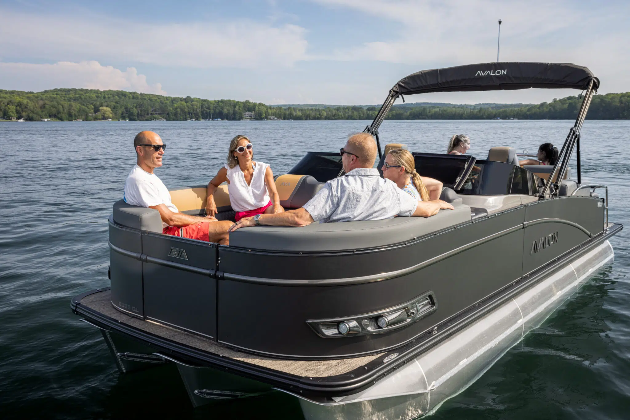 Four people are sitting and chatting on a Catalina Quad Lounger Shift Windshield pontoon boat in calm lake waters. The boat's canopy is partially extended, and green forested hills line the distant shore under a clear blue sky.