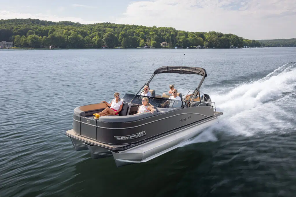 The Catalina Quad Lounger Shift Windshield glides smoothly across the tranquil lake as four passengers unwind and savor the sunlit day. Vibrant green trees border the remote shoreline beneath a sky dotted with clouds.