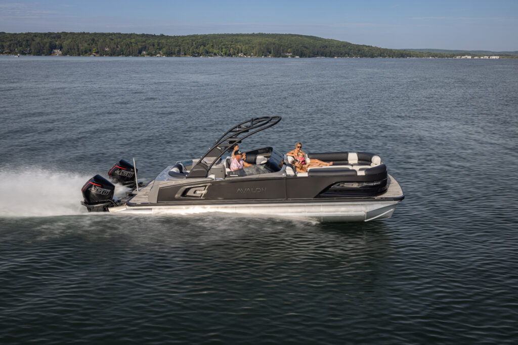 A sleek pontoon boat with multiple people aboard speeds across a calm body of water. The boat has a modern design and powerful engines, with a forested shoreline visible in the background under a clear blue sky.