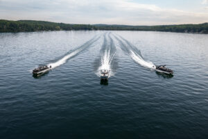 Three motorboats speed across a vast, calm lake, leaving white trails in the water. The scene is surrounded by a lush, green forest under a partly cloudy sky. The boats are evenly spaced and create a dynamic, symmetrical pattern on the water.