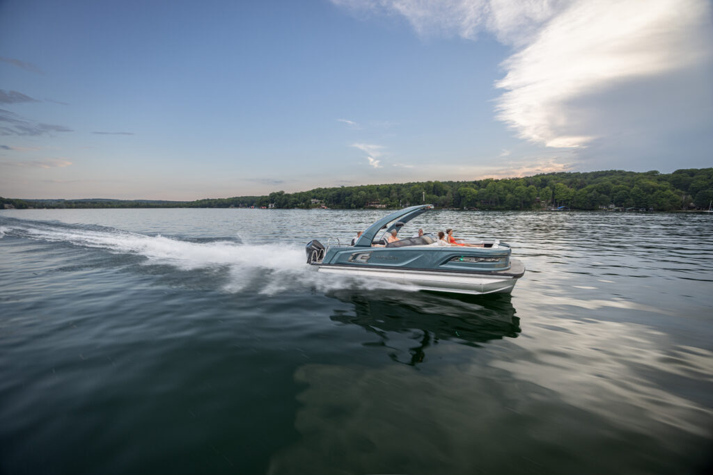 A speedboat cruises across a serene lake under a blue sky with scattered clouds. The boat creates a white wake as it moves, with lush green trees visible along the shoreline in the background.