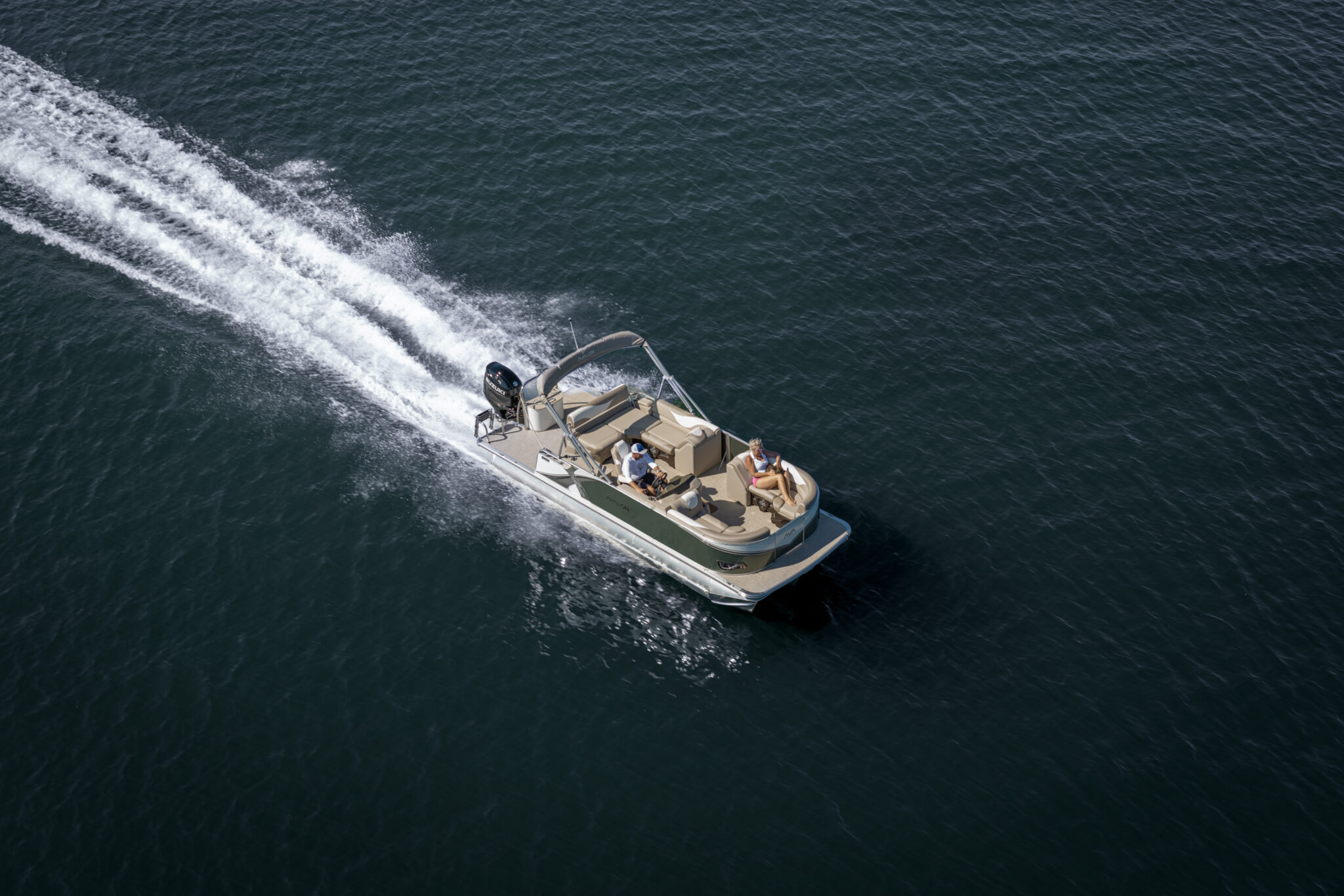 Aerial view of a motorboat speeding across dark blue water with two people on board. The boat creates a white wake as it moves swiftly, set against the vast expanse of the lake or sea.