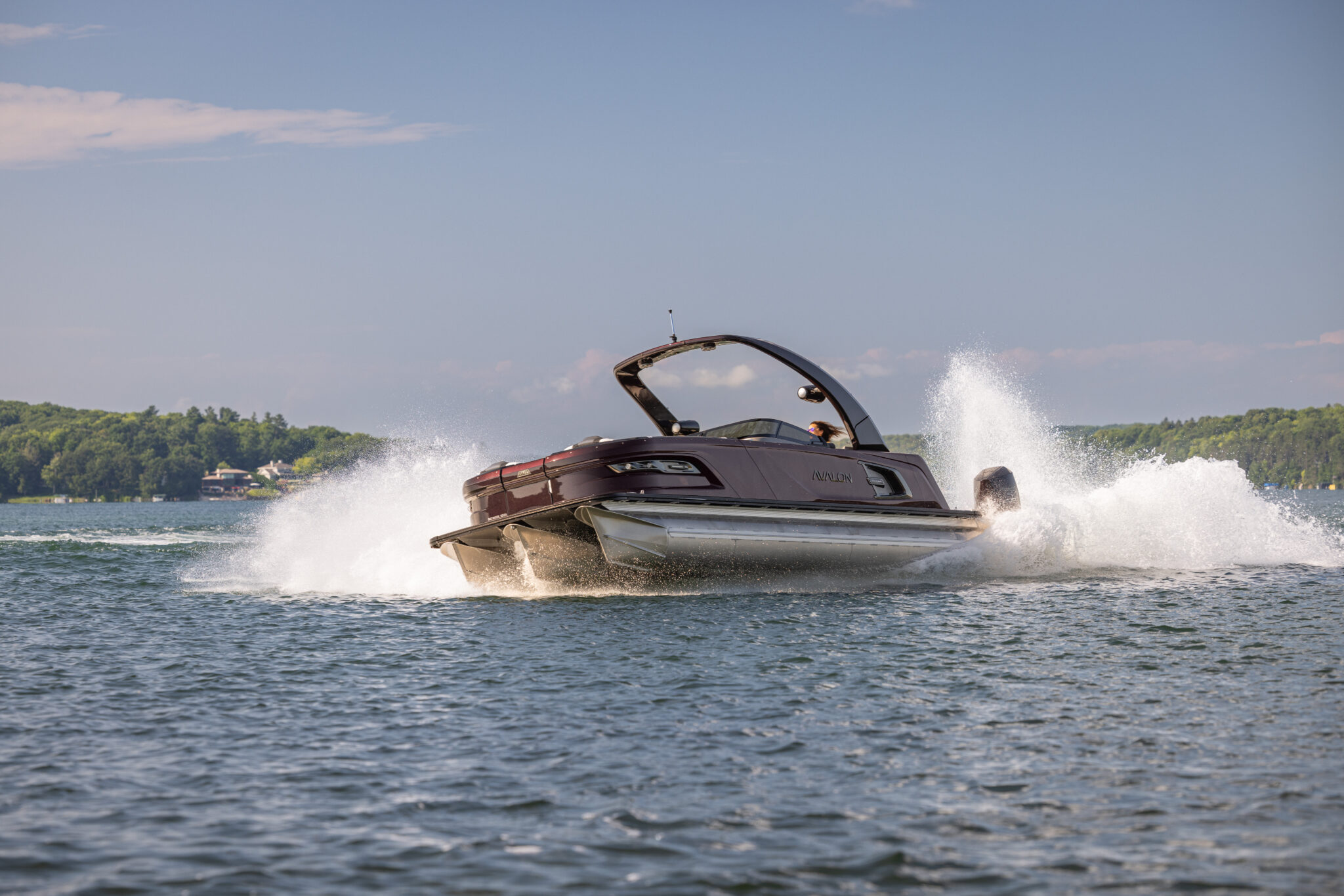 A sleek motorboat speeding on a lake, creating a large splash behind it. The sky is clear with a few clouds, and lush green trees line the distant shore.