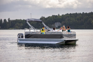 A pontoon boat with several people on board is cruising on a calm lake. One person is steering, while others are sitting and enjoying drinks. The background features a wooded shoreline and a cloudy sky.