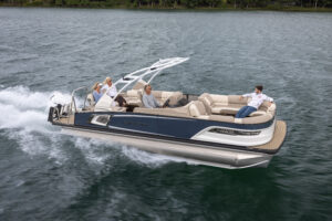 A pontoon boat speeds across a lake, carrying five people. The boat features a sleek design with a white and blue color scheme. Passengers are seated comfortably, enjoying the ride amid lush green trees lining the water's edge.