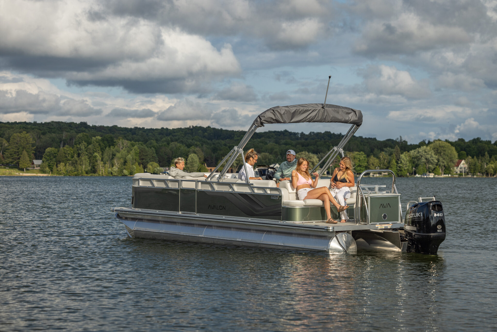 A group of five people relaxes on a Venture 85 Cruise Rear Bench pontoon boat drifting in the middle of a calm lake. The sky is partly cloudy, with lush green trees and distant hills forming the scenic background.