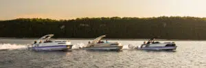 Three motorboats speed across a calm lake under a clear sky, their sleek designs contrasting the pontoon boat's weightier look. Visible passengers enjoy the ride as the forested shoreline mirrors the warm glow of the sunset.