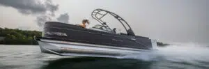 A black pontoon boat, known for its balanced weight distribution, speeds across a lake with water splashing around. A person with long hair sits at the helm under a cloudy sky, and lush green trees line the distant shore.