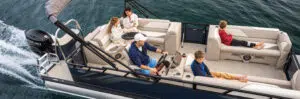 A family enjoys a relaxing day on their pontoon boat. Two adults sit near the helm, while a woman holds a glass and a child sits beside her. Another child lounges at the back, looking at a device. They effortlessly glide through calm water, appreciating how stable their pontoon boat's weight keeps everything.