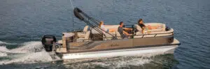 A pontoon boat with four people is gliding across the serene lake, powered by a Mercury outboard motor. The boat features ample seating and a convenient pontoon boat table for added comfort. Small waves ripple as the vessel cuts through the calm waters under a clear sky.