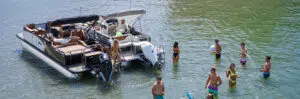 A group of people enjoy a sunny day in a shallow body of water near two pontoon boats. Some are wading and chatting, while others relax on the deck. The lively scene, with its casual conversations and greenery reflected in the water, captures the essence of carefree boating moments.