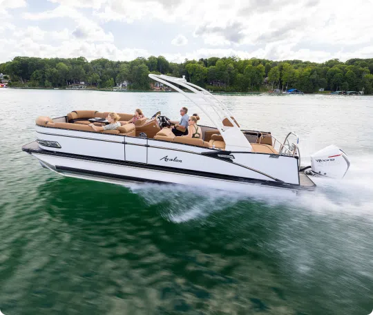 A sleek, white pontoon boat from a top brand speeds across the lake. Several people are seated comfortably on board, enjoying the sunny day. The water is calm, and lush green trees line the distant shore under a partly cloudy sky.