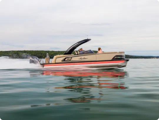 A sleek, modern pontoon boat from top brands speeds across a calm lake, leaving a trail of water in its wake. The boat boasts an arching design with a few people on board. The sky is overcast, and trees line the distant shoreline.