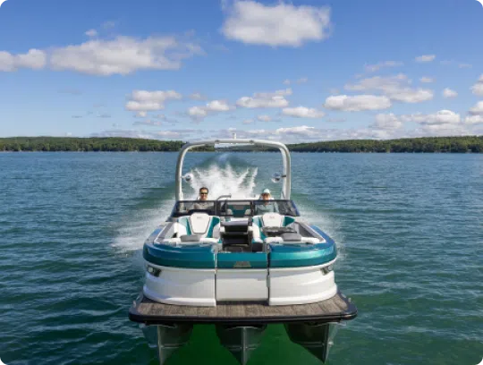 A popular pontoon boat speeds across the lake, leaving a trail of white water. Two people are seated at the front, enjoying the ride. The sky is blue with scattered clouds, and a forested shoreline is visible in the background, epitomizing why top pontoon boat brands are so beloved.