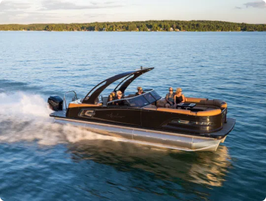 A sleek black pontoon boat from a top brand cruises through a large body of water with four people on board, surrounded by calm blue water and set against a backdrop of dense green forest under a partly cloudy sky.