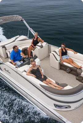 Aerial view of four people relaxing on a white motorboat in open water. The boat, comparable to a sleek pontoon boat, cruises swiftly, leaving a visible trail. The deck features seating and loungers. The passengers are dressed casually, enjoying the sunny day.