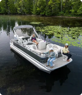 Three people relax on a pontoon boat on a calm, tree-lined lake. The sturdy build supports them effortlessly as two sit at the back fishing, while the third enjoys the shade under a canopy at the front. Lily pads float nearby.