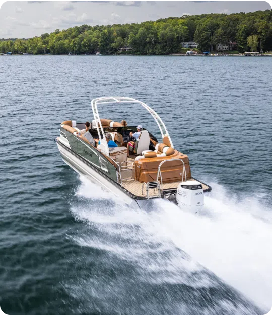 A motorboat speeds across a tranquil lake, leaving a foamy wake as it navigates like a pontoon boat under the influence of weight distribution. The boat features brown seating and a white canopy, while dense green trees line the distant shoreline under a partly cloudy sky.