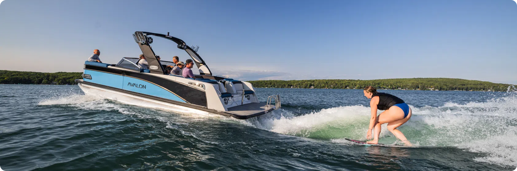 A woman wake surfing on a lake near a blue and white boat with several passengers, enjoying the day around a pontoon boat adorned with a table. The sun is shining, and the landscape boasts a forested shoreline under a clear blue sky.