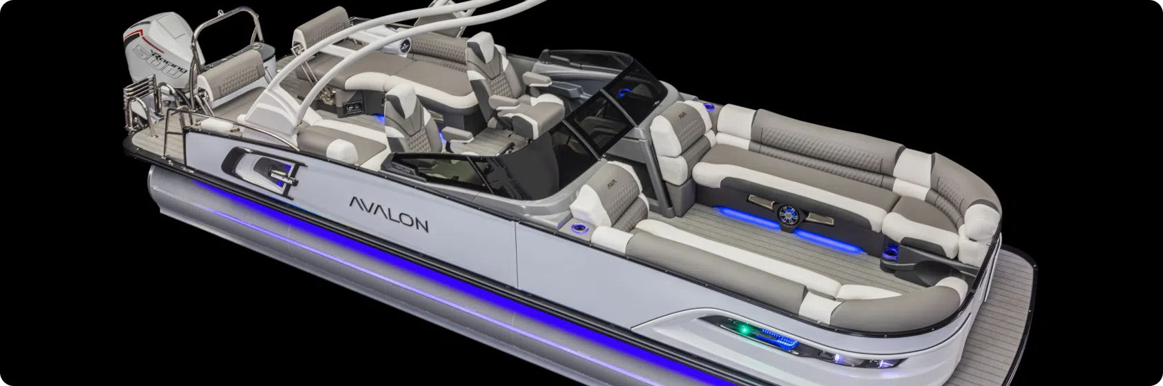 A luxurious pontoon boat with white and gray seating and an Avalon logo on the side, this vessel features multiple seating areas with a sleek pontoon boat table, a control console, and blue LED lights underneath. The motor stands prominently at the rear against a black background.