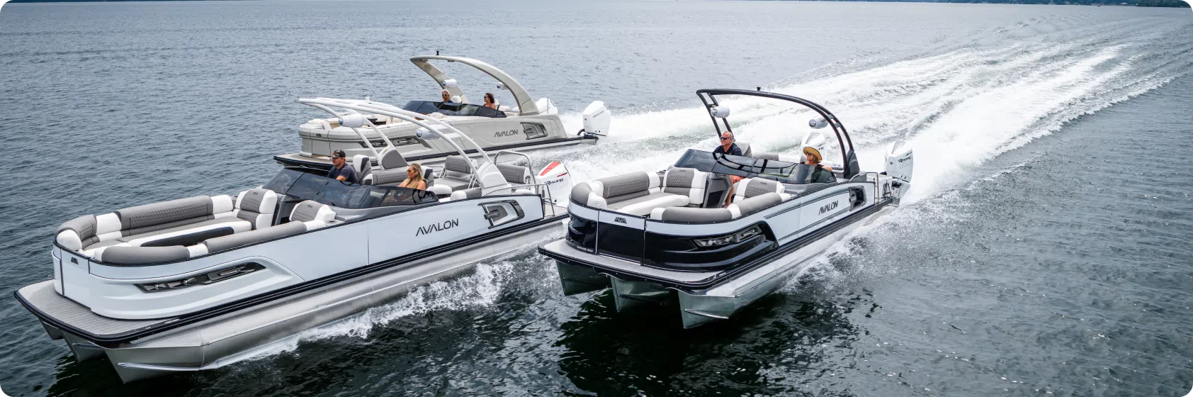 Two luxury pontoon boats speed across a large body of water, their modern designs featuring sleek seating and elegant pontoon boat tables. The arching frames glide side by side, creating waves as they move swiftly under a cloudy sky.