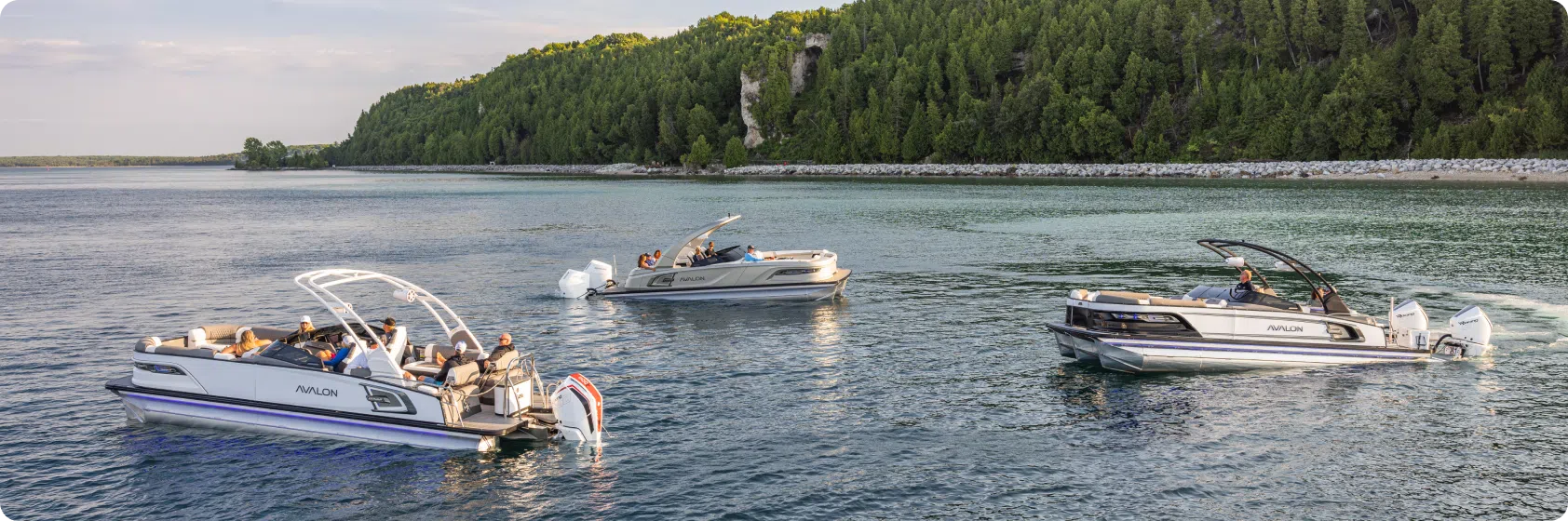 Three luxury pontoon boats with passengers, complete with white engines and elegant tables, cruise along a calm lake. The backdrop features lush green trees and a rocky shoreline under a partly cloudy sky.