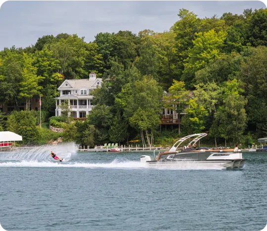 A person is waterskiing on a lake, expertly navigating the chop created by the motorboat's wake. The boat slices through the water with precision. In the background, lakeside houses are cradled by lush green trees under a clear sky, adding to the serene aquatic tableau.