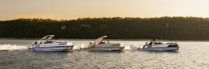 Three sleek motorboats speed across a calm lake, leaving white trails behind them. The boats have white hulls and feature curved windshields. A lush green forest and a warm, golden sky provide a scenic backdrop.