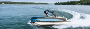 A sleek new blue pontoon boat with "Avalon" on the side glides quickly across a clear lake, creating waves. Lush green trees line the distant shore under a partly cloudy sky.