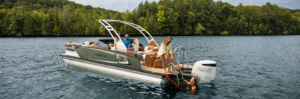 A family enjoys a day on a motorboat in a calm lake. One person stands on the deck, two sit at the helm under a canopy, and a swimmer holds onto the boat's ladder. Dense green trees line the shoreline in the background.