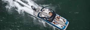 A top view of a motorboat speeding across the water. Several passengers relax on the deck chairs under the sun. The water is green, and the boat creates a white wake as it moves forward.