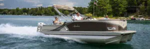 A pontoon boat with two men and a woman glides swiftly across the lake surrounded by trees, leaving a trail of water. The sky is partly cloudy as an American flag flutters proudly on this vessel crafted by one of the top pontoon boat brands.