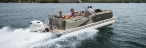 A new pontoon boat with four people is speeding across a lake. The passengers are seated, enjoying the ride, as the boat creates a trail of white water behind it. The background features a shoreline with trees.