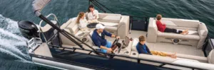 A family is enjoying a ride on a pontoon boat. The man is steering, while the woman and children relax on the seats, some with drinks and snacks. The boat is moving across calm, dark green water.