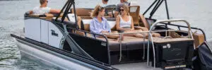 A group of people relaxes on a sleek pontoon boat. Two women in casual attire chat while seated on the deck, and two men are at the helm discussing their favorite pontoon boat brands. The boat is on calm water, creating a sunny and peaceful scene.