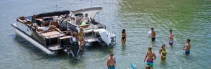 A group of people stands in shallow water near a pontoon boat on a sunny day. Some are wearing swimwear, and two individuals are on the boat. The water is clear, and the scene is relaxed and recreational.