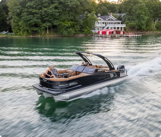 A pontoon boat with several passengers glides across a lake, leaving a wake behind. The boat features a sleek design and tan seating. In the background, a large lakeside house with red and white accents is surrounded by lush trees.