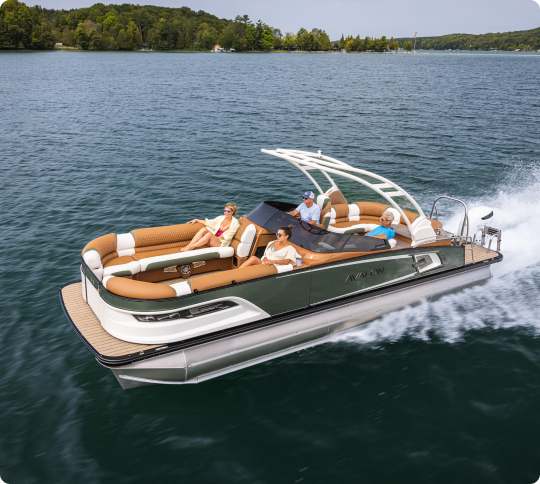 A pontoon boat with four people on a lake. The boat features a modern design with brown seating and a white canopy. It speeds across the water, creating a wake behind it. Lush green trees line the distant shore under a clear sky.