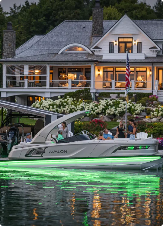 A boat with green underglow lights is floating near a large, well-lit house by a lake. Several people are seated on the boat, and the house has a stone chimney and garden with white flowers. An American flag is on display.