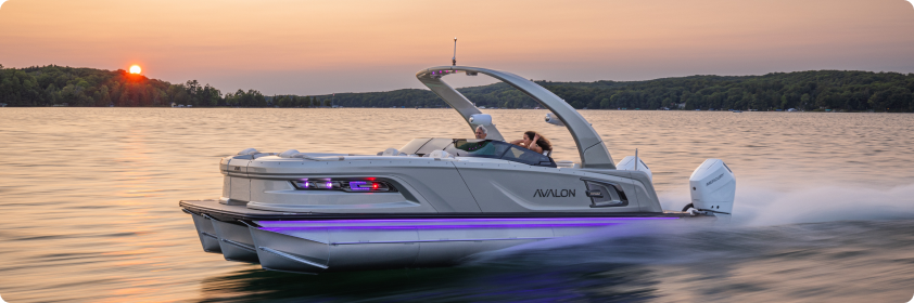 A sleek pontoon boat speeds across a tranquil lake at sunset, leaving a trail of frothy water. The warm glow of the sun is setting behind tree-lined shores. The boat has seating and modern design features, and two people are visible enjoying the ride.