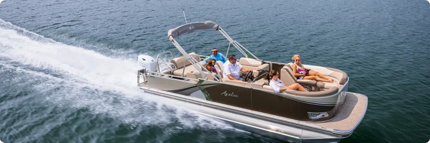 A pontoon boat cruising on a sunny day with five people relaxing on board. The boat creates a small wake as it moves through the calm water. The passengers appear to be enjoying the ride and the scenic view.