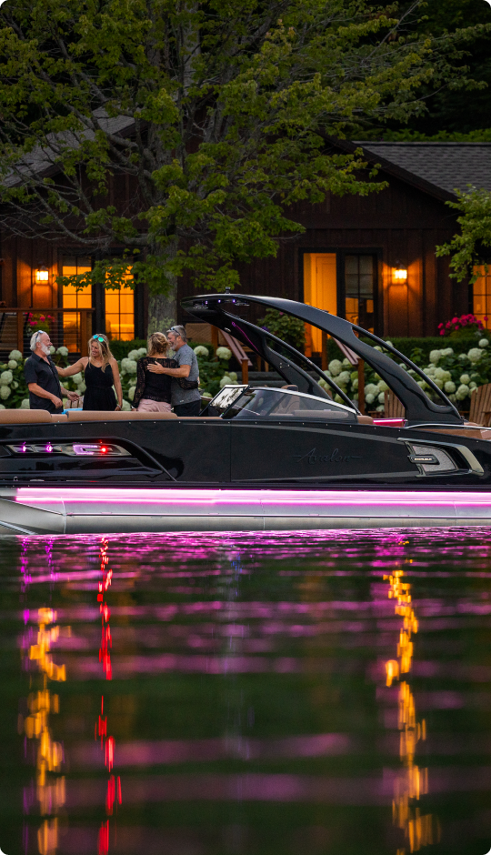 A sleek boat with purple lights is docked on a calm lake. Four people are standing on the deck, talking. A cozy wooden house with warm lit windows and lush trees are visible in the background. The water reflects colorful lights.