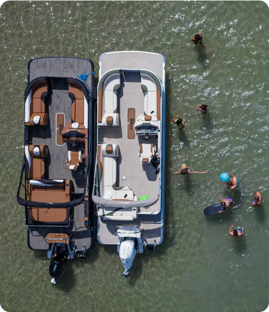 Aerial view of two pontoon boats anchored closely in clear shallow water. Several people are swimming around the boats, some wearing life vests and holding paddles, creating a lively, recreational scene.