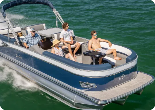 A pontoon boat glides over the water. Four people are on board: one at the steering console, two seated under shade, and one lounging shirtless on a recliner. The water is a deep green, and the boat has blue and white accents.