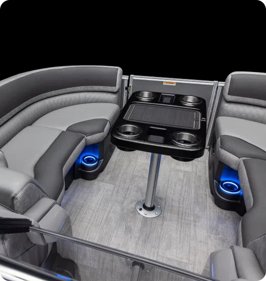 Interior of a luxury pontoon boat featuring curved gray seating with cup holders and blue LED lighting. A rectangular table with built-in cup holders is centered on a carpeted floor.