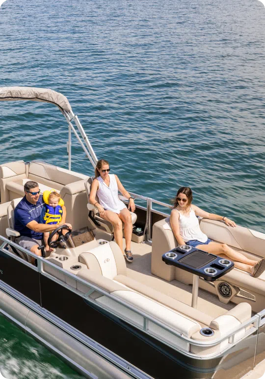 A group of four people, including a child in a life jacket, are relaxing on a pontoon boat on a sunny day. The boat is cruising through calm waters, and the adults are seated comfortably while enjoying the open air.