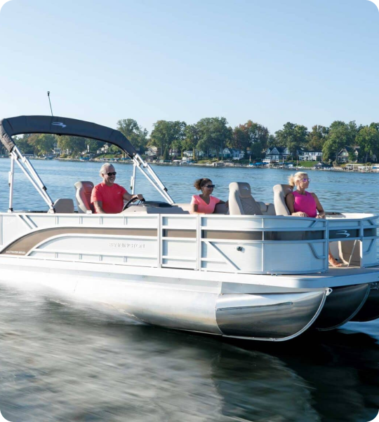 Three people are seated on a silver pontoon boat speeding across a lake. The boat has a canopy, and the water is calm. Trees and houses line the distant shoreline under a clear blue sky.