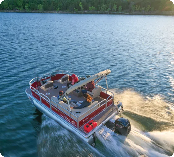 A red pontoon boat with a canopy glides across a calm lake, leaving a wake behind. Two people are seated on the boat, enjoying the scenic surroundings, with trees lining the distant shore under a clear sky.